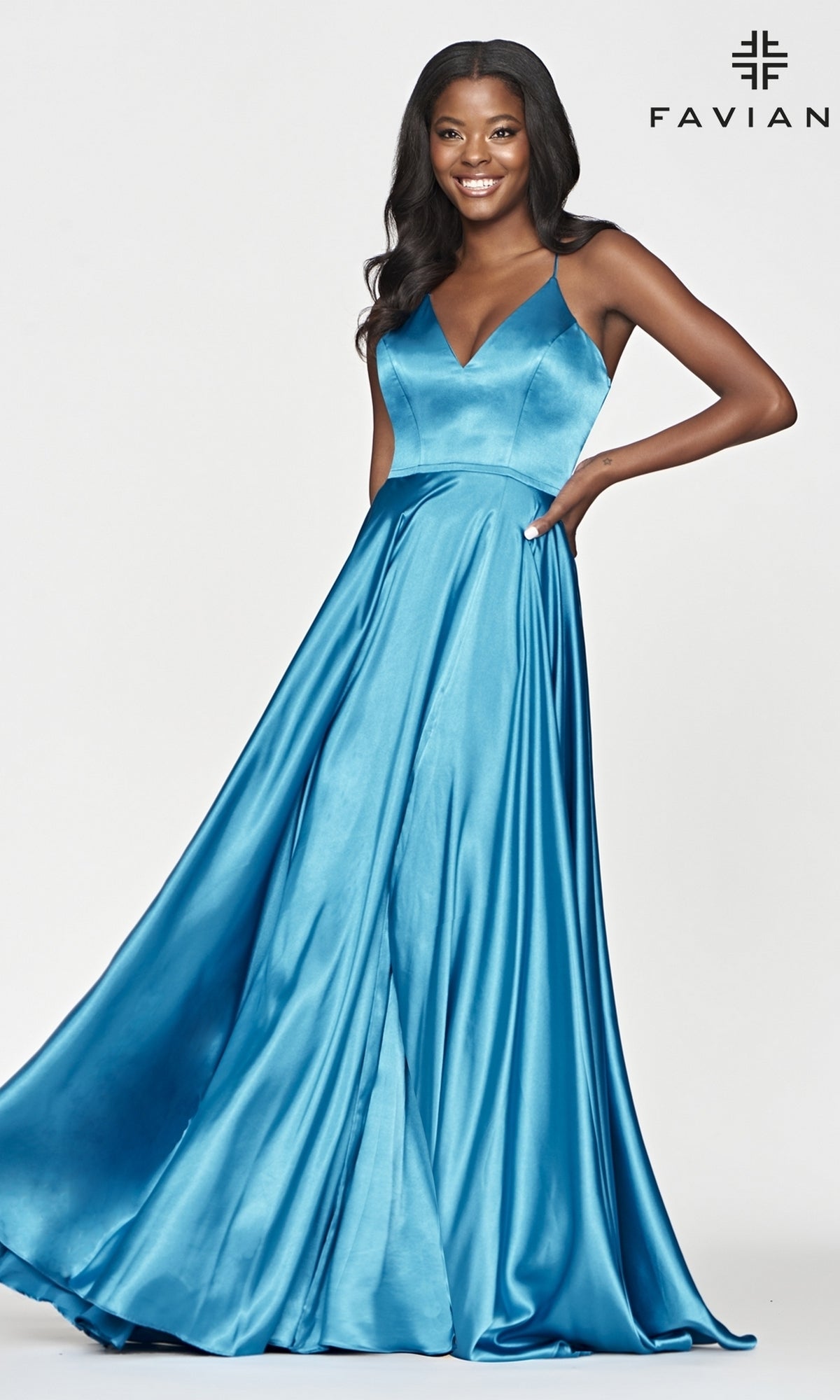 A woman poses smiling with one hand on her hip in the Long Faviana Prom Dress with Back Cut Out, a shiny blue gown featuring a V-neck and thin straps, exuding elegance against a plain white background.